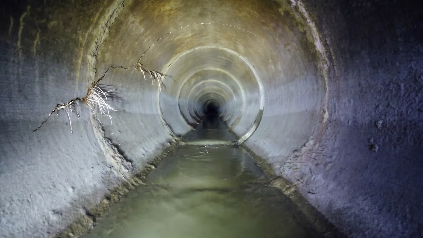 Interior CCTV camera view down a sewer lateral pipe showing a wall crack with tree roots intruding into the line, above flowing wastewater